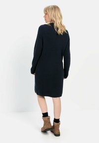 Dunkelblaues geripptes Strickkleid mit langen Ärmeln, gerippter Textur, Seitenschlitzen, getragen mit braunen Ankle Boots und schwarzen Socken.