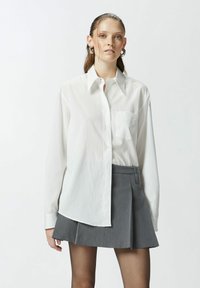 Camicia a maniche lunghe bianca a righe con chiusura a bottoni, dotata di una tasca sul petto. Abbinata a una mini gonna a pieghe grigia.
