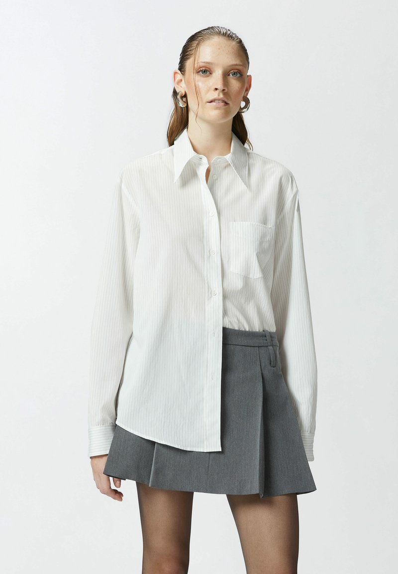 Camisa blanca de manga larga a rayas con botones en la parte delantera, que cuenta con un bolsillo en el pecho. Combinada con una falda corta gris con pliegues.