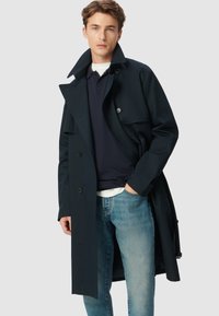 Cappotto trench blu navy con design doppiopetto, ampio colletto e spalline. Indossato sopra un polo blu navy, abbinato a jeans azzurri.
