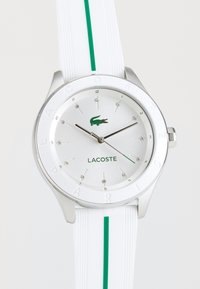 Lacoste laikrodis su sidabriniu ciferblatu, balta silikono juosta su žalia juostele ir nerūdijančio plieno korpusu. Apima kristalines žymekles ir logotipą.