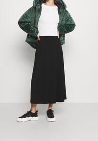Veste matelassée verte, haut blanc à manches longues côtelées, jupe midi noire, et baskets épaisses noires et blanches. Silhouette moderne et structurée.