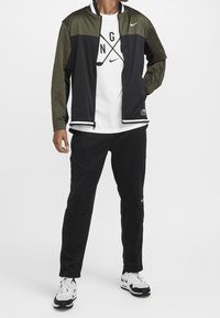 Sportowy strój Nike składający się z dwukolorowej kurtki w ciemnej zieleni i czerni z zamkiem błyskawicznym, w połączeniu z czarnymi spodniami i białymi butami sportowymi.