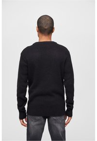 Un suéter negro de punto con mangas largas, puños acanalados y dobladillo. Llevado con jeans grises, visto desde la parte posterior contra un fondo simple.