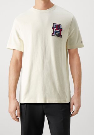 Man wearing a cream short-sleeve t-shirt with a large multicolor "TH" logo on the chest and small logo on the sleeve.