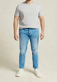 Homme portant un t-shirt à manches courtes gris clair, un jean slim bleu clair et des baskets blanches, debout devant un fond beige uni.