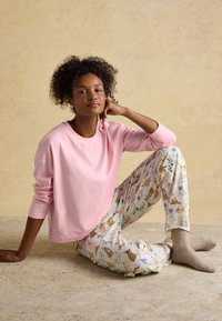 Woman with curly hair wearing pink long-sleeve shirt and dog-patterned pajama pants sits relaxed on beige floor and wall background.