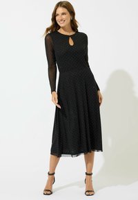 Abito lungo nero a maniche lunghe con scollatura a chiave, motivo a pois e gonna fluida, caratterizzato da un materiale trasparente sulle maniche e sull'orlo.