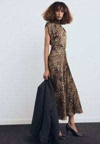 Mujer con cabello rizado vistiendo un vestido sin mangas de estampado de leopardo y tacones negros, sosteniendo un abrigo oscuro contra una pared blanca lisa.