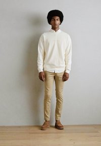 Creme sweatshirt med ribbede manchetter, beige chinos og lysebrune sko. Modellen har en rund afrofrisure og står foran en neutral baggrund.