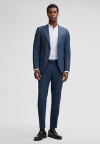 Maßanzug aus dunkelblauem Stoff mit subtilen Mustern. Ausgestattet mit einem Ein-Knopf-Blazer und schmal geschnittenen Hosen, kombiniert mit einem weißen Hemd.