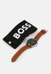 Orologio con cinturino in pelle marrone, quadrante nero, cassa in argento e dettagli bianchi. Include una borsa di stoffa nera con la scritta "BOSS" in caratteri bianchi in grassetto.