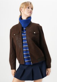 Moteris vilki rudą zip-up striukę ant mėlynos dryžuotos polo megztinio ir tamsiai mėlynos klostuotos sijono, žiūri į kairę prieš baltą foną.