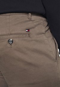 Brązowe bawełniane chinosy z tylną kieszenią, z małym haftowanym logo w czerwonym, białym i niebieskim kolorze oraz czarnym zapięciem na guzik.