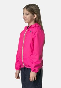 Giacca leggera di un brillante colore rosa con cappuccio, polsini elasticizzati e zip frontale. Presenta un piccolo logo sul petto. Abbinata a pantaloni scuri.