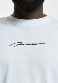 Bílý bavlněný tričko s černým vyšívaným logem "Rocawear" v kurzívě přes hrudník. Kulatý výstřih, měkká textura.