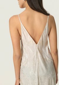 Woman with long dark hair wearing a sleeveless light beige dress with thin straps and a deep V-back, standing against a plain light background.