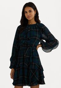 Grønn rutete kjole med volangermed og hemline, innsnevret midje og bånd i halsen. Laget av et lett, strukturet stoff.