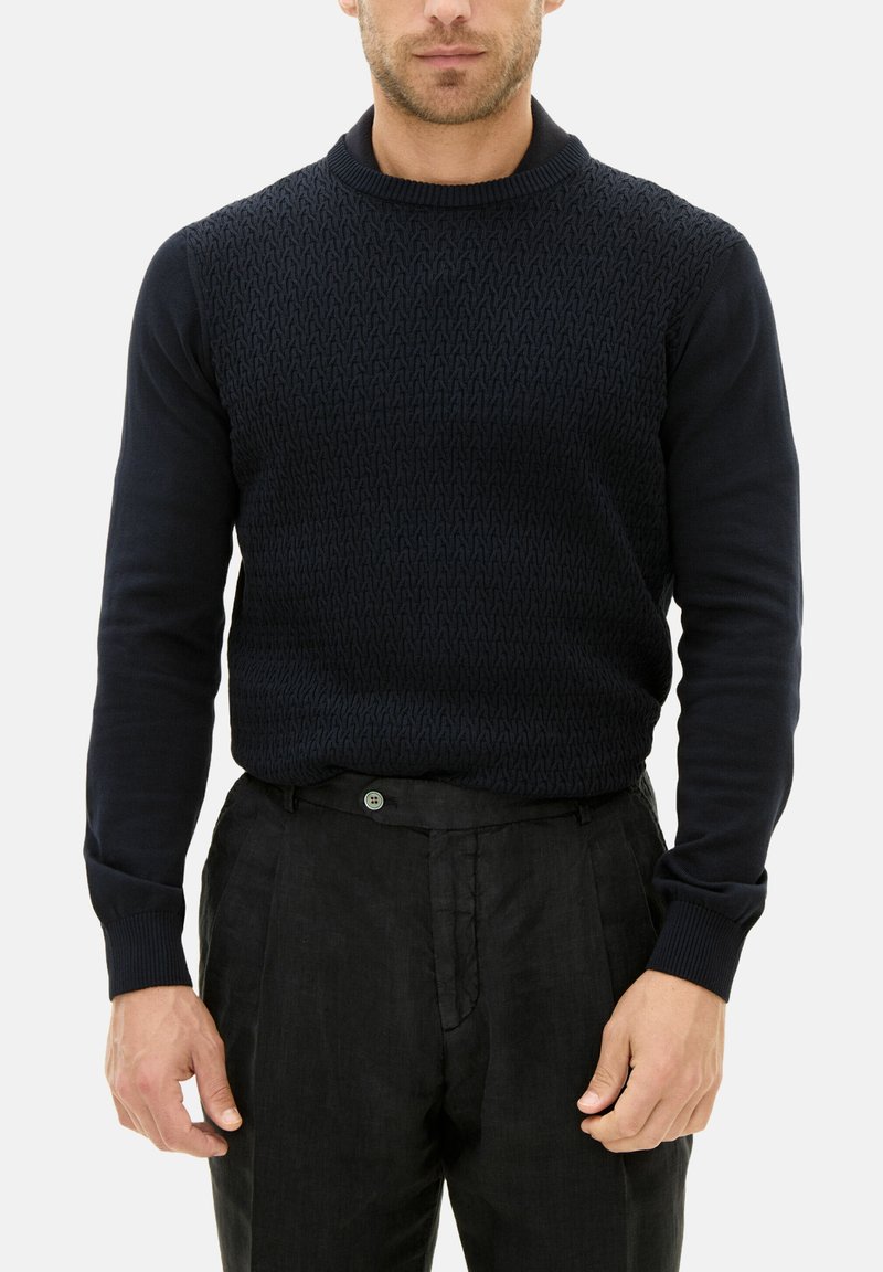 Dunkelblauer Strickpullover mit einem strukturierten Muster auf der Brust und glatten Ärmeln, kombiniert mit schwarzen Hosen, die einen grünen Knopf haben.