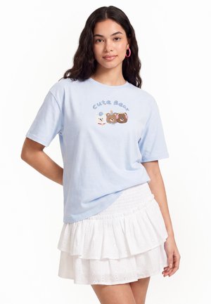 Młoda kobieta z długimi ciemnymi włosami, ubrana w jasnoniebieski t-shirt z trzema twarzami niedźwiedzi i tekstem "Cute Bear", w połączeniu z białą spódnicą warstwową.