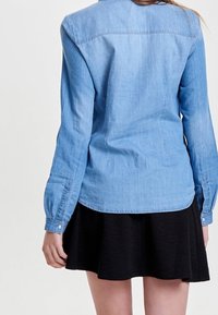 Camicia di jeans azzurro chiaro con maniche lunghe, polsini abbottonati e orlo curvo, abbinata a una gonna nera a ruota. Vista posteriore.