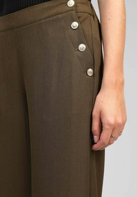 Pantalons marron sur mesure présentant une texture lisse, des accents de boutons sur les côtés et une taille ajustée. Les boutons sont métalliques et décoratifs.