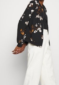Chaqueta bomber negra con estampado floral en beige y marrón. Cuenta con cierre de cremallera y mangas ampias, llevada sobre una camiseta negra y pantalones blancos.