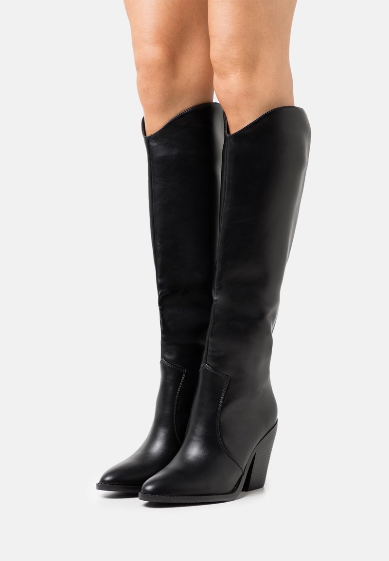 Bottes en cuir noir montantes jusqu'aux genoux, à bouts pointus et talons blocs, portées sur des jambes nues sur un fond clair uni.