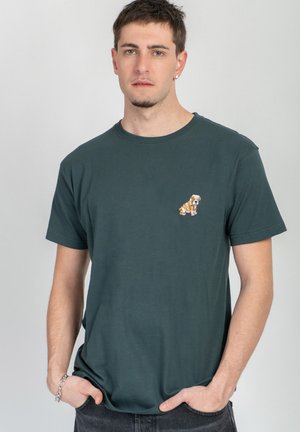 Giovane uomo con capelli corti e scuri che indossa una maglietta verde scuro con un piccolo bulldog ricamato sul petto, mani nelle tasche, sfondo neutro.