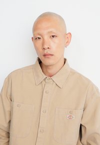 Camisa marrón claro de botones con dos bolsillos en el pecho, hecha de una tela texturizada. Presenta un parche de logo, diseño con collar y detalles de costura visibles.