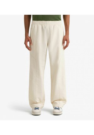 Persona che indossa pantaloni larghi color bianco sporco con vita elastica e scarpe da ginnastica bianche con lacci blu, in piedi davanti a uno sfondo semplice.