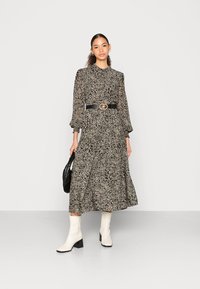 Vestido longo com padrão de animal em preto e bege, com mangas bufantes, cinto preto ajustado e botas brancas de salto. A modelo segura uma bolsa preta.