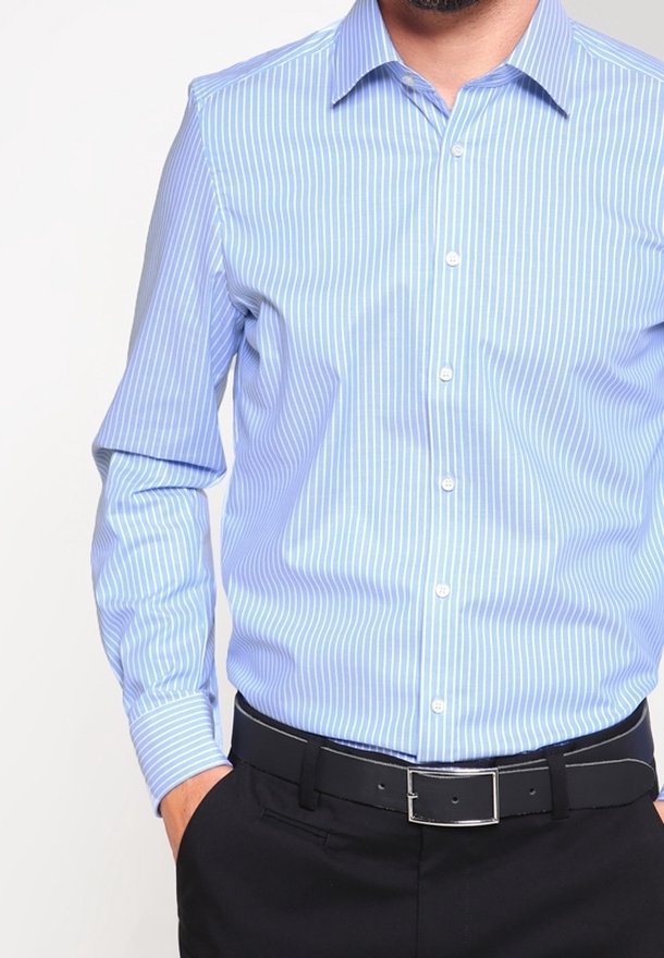 Homme portant une chemise à rayures verticales bleu clair et blanc rentrée dans un pantalon noir avec une ceinture noire, main dans la poche.