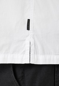 Camisa blanca de botones con una etiqueta negra, tela texturizada y costuras visibles, cubriendo parcialmente pantalones negros.