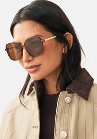 Lunettes de soleil avec une teinte brune, une monture carrée et des accents métalliques dorés. Portées avec une veste beige dotée d'un col en velours côtelé.