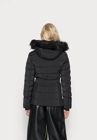 Doudoune noire avec capuche en fausse fourrure, design matelassé, taille ceinturée et accents côtelés sur les côtés. Portée sur un pantalon noir à jambes larges.