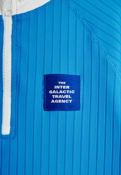 Μπλε υφασμάτινη ύφανση με λευκό φερμουάρ και ραμμένο έμβλημα που αναγράφει «THE INTER GALACTIC TRAVEL AGENCY» με λευκό κείμενο.