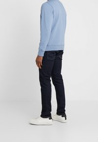Felpa azzurra chiara con polsini e orlo a costine, abbinata a jeans denim scuri. Sneakers bianche con dettagli neri sul tallone, viste dal retro.