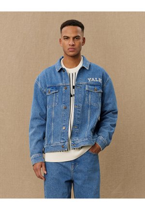 Denim jakna v svetlo modri barvi, z dvema žepoma na prsih, gumbi za zapiranje in izvezenim napisom "Yale" na levi strani. Tekstura tkanine.