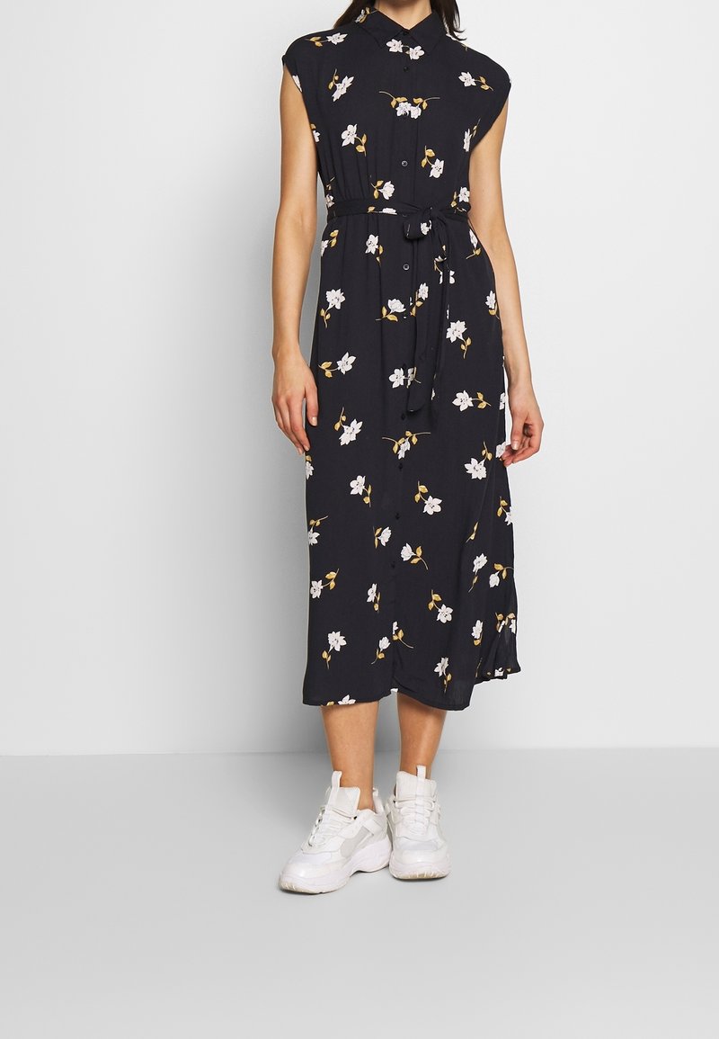 Marineblaues Blumenhemdkleid mit kurzen Ärmeln, Knopfschluss und einem Bindegürtel. Aus einem weichen, fließenden Stoff mit weißen Blumen und goldenen Blättern.