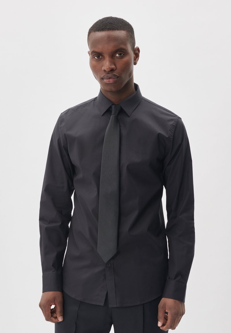 Camisa de vestir negra en un diseño ajustado, con collar clásico y cierre de botones, combinada con una corbata negra a juego, de textura suave.