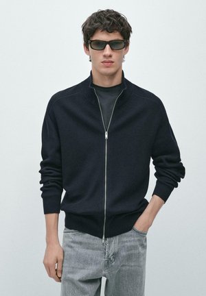 Jeune homme portant un pull zippé noir, un T-shirt noir, des lunettes de soleil et un jean gris clair, debout avec une main dans la poche.