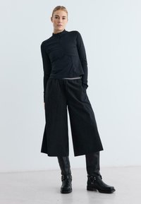 Frau trägt einen schwarzen Langarmzipper, weit geschnittene, knöchellange Hosen und schwarze Kniefallstiefel mit Schnalle, steht vor einem schlichten weißen Hintergrund.