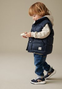 Gilet imbottito blu navy con cappuccio sopra una felpa grigia chiara, abbinato a jeans blu e sneakers navy con dettagli marroni. Il bambino tiene un piccolo oggetto.