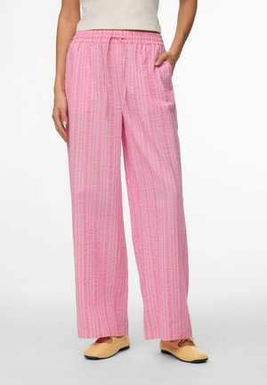 Kvinde iført løse lyserøde bukser med striber, elastik i taljen og snøre, kombineret med beige flade sko, stående med den ene hånd i lommen.