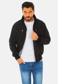 Giubbotto bomber nero con tasche con zip, tessuto morbido, polsini e colletto a coste; indossato sopra un maglione bianco e jeans blu.