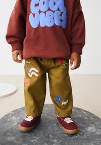 Sweatshirt marron avec le texte bleu "COOL VIBES", associé à un pantalon vert olive avec des patches colorés et une paire de baskets bordeaux.