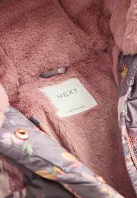 Doublure rose duveteuse de veste avec une étiquette blanche portant l'inscription « NEXT », montrant une partie d'une coque extérieure à motif floral ainsi qu'une fermeture éclair et des boutons-pression en cuivre.