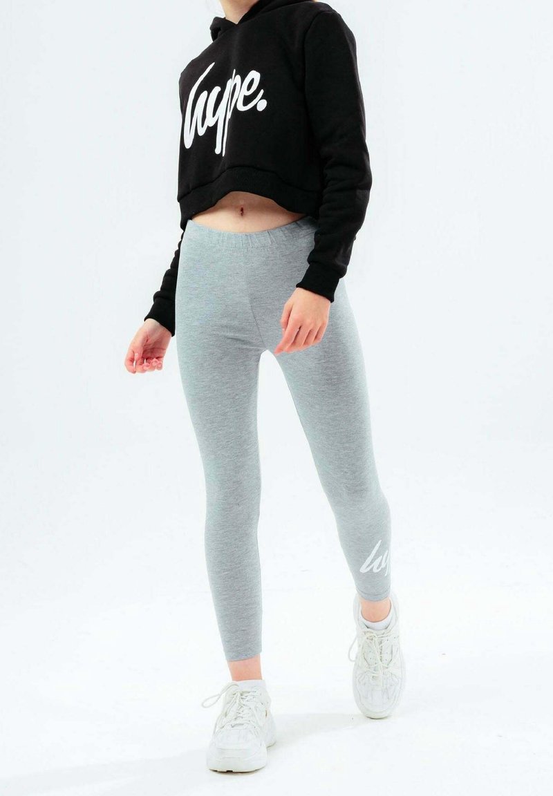 Personne portant un sweat à capuche court noir avec le texte « Hype. », des leggings gris clair et des baskets blanches marchant sur un fond blanc.