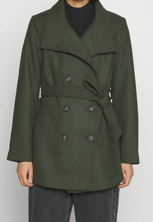 Personne portant un manteau ceinturé croisé vert foncé à larges revers, par-dessus un haut noir et un pantalon gris foncé, debout devant un fond uni.